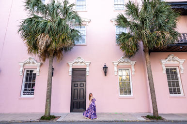 Person in a purple floral maxi dress and sunglasses poses on a sidewalk in front of a pastel pink historic building with ornate white window trim, a black door and lantern, flanked by tall palm trees — colorful palm-lined street scene.