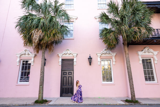 Person in a purple floral maxi dress and sunglasses poses on a sidewalk in front of a pastel pink historic building with ornate white window trim, a black door and lantern, flanked by tall palm trees — colorful palm-lined street scene.