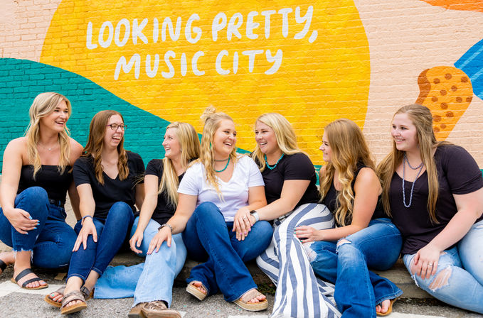Seven women laughing and sitting on a sidewalk in front of a colorful “Looking Pretty, Music City” mural, casual jeans and sandals against a vibrant Nashville street-art backdrop.