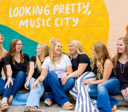 Seven women laughing and sitting on a sidewalk in front of a colorful “Looking Pretty, Music City” mural, casual jeans and sandals against a vibrant Nashville street-art backdrop.