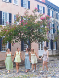 Five friends in summer dresses holding hands and laughing as they stroll down a historic cobblestone street lined with pastel row houses and a blooming pink crepe myrtle tree.