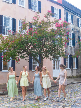 Five friends in summer dresses holding hands and laughing as they stroll down a historic cobblestone street lined with pastel row houses and a blooming pink crepe myrtle tree.