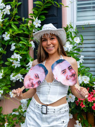 Smiling woman in a white beaded cowboy hat and matching outfit holding two large smiling face cutout props in front of a lush white-flowered garden backdrop