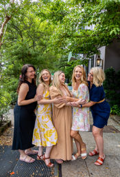 Five women laughing and hugging on a tree-lined sidewalk, wearing colorful summer dresses and sandals in a cheerful outdoor group portrait