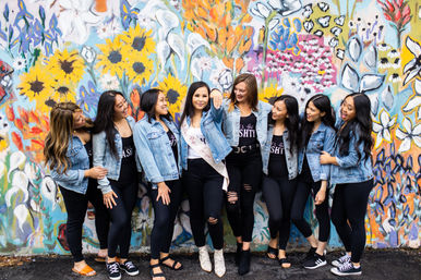 Eight friends in denim jackets and black outfits strike a playful pose and laugh in front of a colorful street floral mural; the central woman wears a sash and white boots.