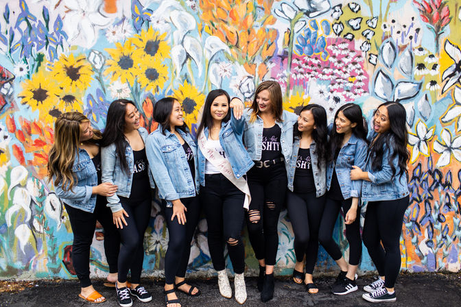 Eight friends in denim jackets and black outfits strike a playful pose and laugh in front of a colorful street floral mural; the central woman wears a sash and white boots.