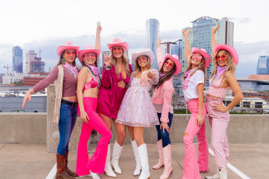 Eight women in pink cowgirl hats, boots and sunglasses striking celebratory poses on a rooftop with a downtown skyline backdrop, bright and playful group photo.