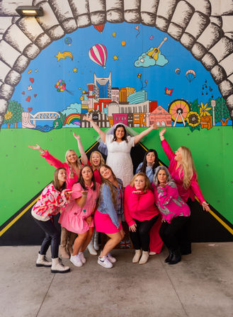 Bride in a white dress and veil with friends in pink posing and blowing kisses in front of a vibrant downtown-style mural featuring a colorful city skyline, hot air balloons, ferris wheel and rainbow.