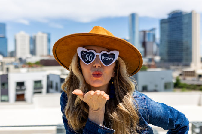 Woman on an urban rooftop blowing a kiss, wearing a tan wide-brim hat, denim jacket and heart-shaped sunglasses reading "I'm 50", city skyline in the background