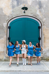 Five friends in coordinated blue outfits laughing and walking arm-in-arm in front of a large teal arched door set in a weathered stone facade on a city sidewalk.