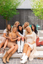 Seven women friends in neutral outfits sitting on city plaza steps by a cascading water wall and trees, smiling and posing in casual summer fashion with boots.
