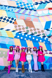 Six women in pink and white outfits with cowboy hats and boots posing and smiling in front of a vibrant geometric polka-dot street mural painted on a brick wall