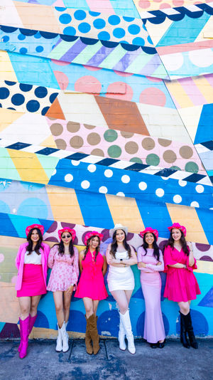 Six women in pink and white outfits with cowboy hats and boots posing and smiling in front of a vibrant geometric polka-dot street mural painted on a brick wall