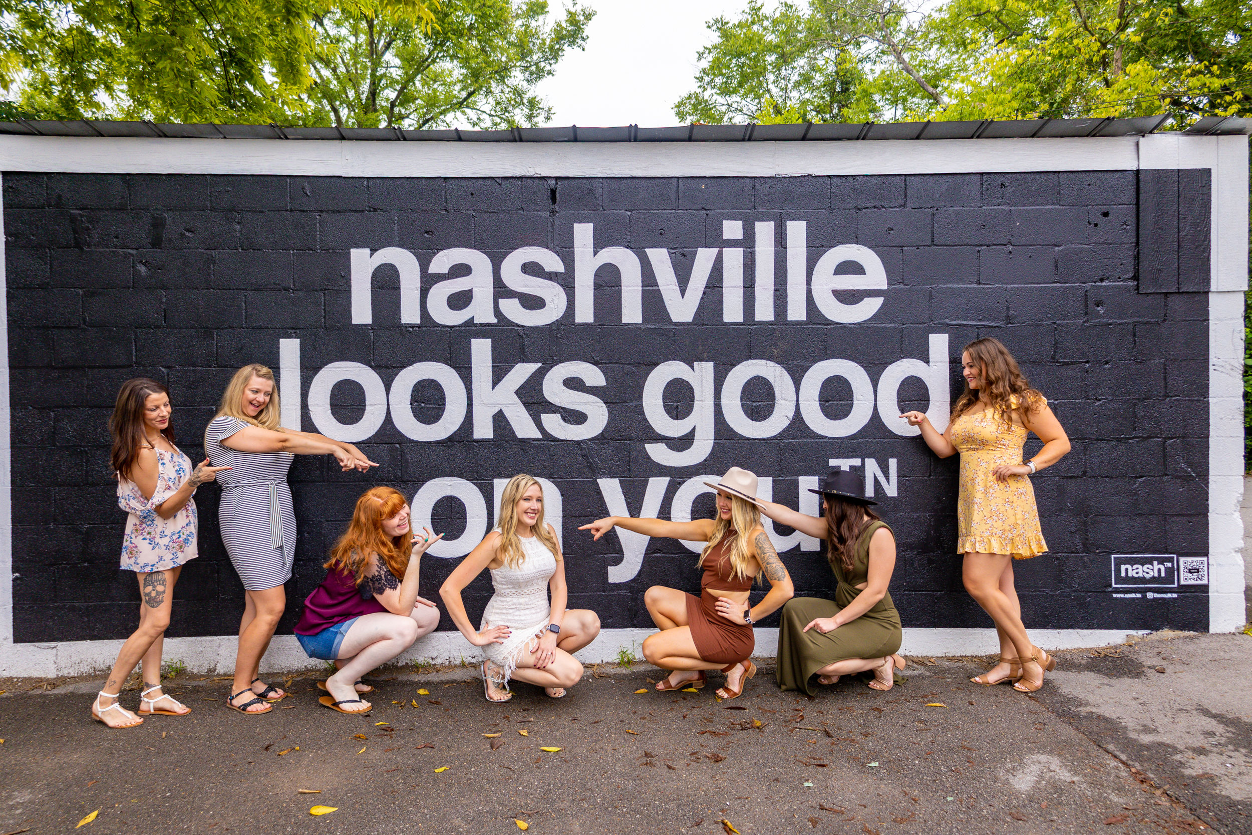 Seven women in summer dresses posing and pointing at a large black mural that reads 'nashville looks good on you' on a tree-lined street.