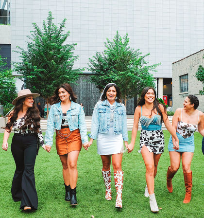 Five smiling women holding hands and walking across a grassy urban plaza with trees and modern buildings, wearing trendy denim jackets, cow-print skirts and boots.