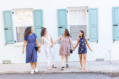 Four friends laughing and holding hands as they stroll past a white building with mint-blue shutters on a sunny sidewalk