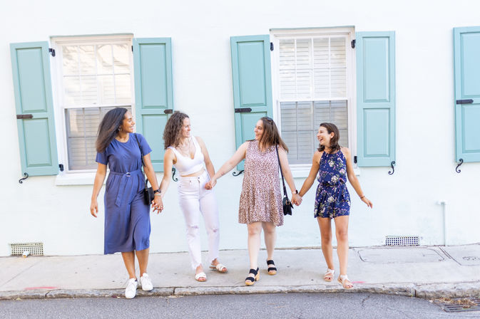 Four friends laughing and holding hands as they stroll past a white building with mint-blue shutters on a sunny sidewalk