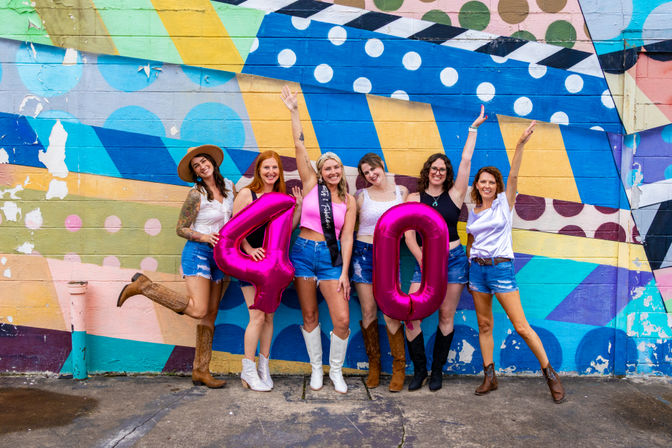Group of six women in denim shorts and cowboy boots celebrating a 40th birthday, holding pink metallic '4' and '0' balloons in front of a vibrant geometric street-art mural