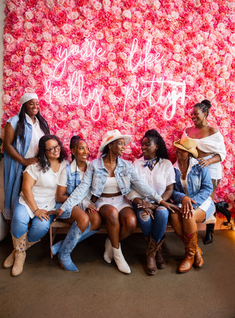Group of seven smiling women in white and denim outfits with boots and hats, seated on a bench in front of a vibrant pink rose floral wall with a neon cursive sign — bright, celebratory group photo.