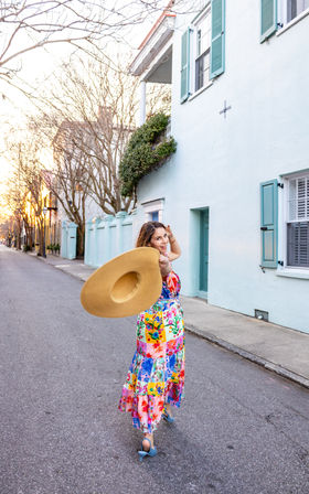 Woman in a colorful floral maxi dress twirling a wide-brim straw hat while walking down a quiet street lined with pastel row houses, shuttered windows, and leafless trees at sunset.