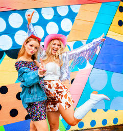 Two friends striking a playful pose in front of a colorful street-art mural — one in a sequin dress and denim jacket flashing a peace sign, the other in a pink cowboy hat, silver fringe jacket, cow-print skirt and white boots kicking a leg.