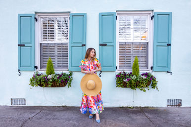 Person in a bright floral dress holding a wide straw sun hat stands against a pastel blue house with teal shutters and lush window boxes of purple and white blooms on a quaint sidewalk.