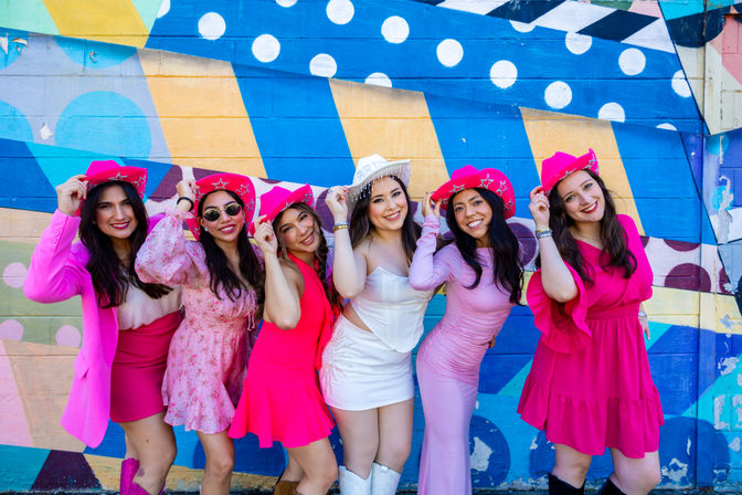 Bachelorette party: six friends in pink outfits and cowboy hats with the bride in a white mini dress, laughing in front of a colorful geometric street-art mural on an urban wall.