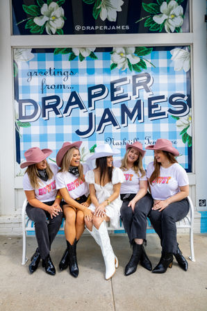Five friends in cowgirl hats and boots laughing on a bench outside a blue gingham floral storefront window display in Nashville, one wearing a white dress and the others in matching 'howdy' tees.