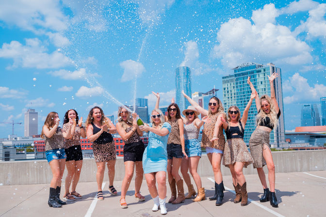 Group of friends celebrating on an urban rooftop parking deck, spraying champagne and cheering under a bright blue downtown skyline.
