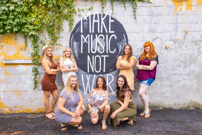 Seven women posing and smiling in front of a “Make Music Not War” mural on a vine-covered urban brick wall, summer outfits and street art in a downtown alley group photo