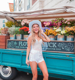 Smiling woman in a white cowgirl hat and sparkly shorts posing by a turquoise flower truck loaded with colorful bouquets on a sunny city street