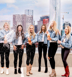 Six women in denim jackets popping champagne and laughing on an urban rooftop with a city skyline backdrop, champagne spray sparkling in the air.