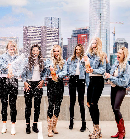 Six women in denim jackets popping champagne and laughing on an urban rooftop with a city skyline backdrop, champagne spray sparkling in the air.