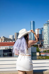 Bride-to-be in white crop top, mini skirt and beaded cowboy hat with veil reading 'bride to be,' posing on a sunny urban rooftop with modern downtown skyline and clear blue sky.