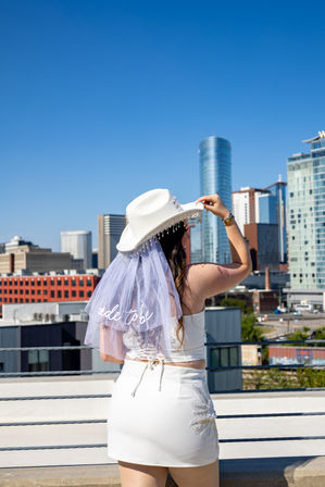 Bride-to-be in white crop top, mini skirt and beaded cowboy hat with veil reading 'bride to be,' posing on a sunny urban rooftop with modern downtown skyline and clear blue sky.