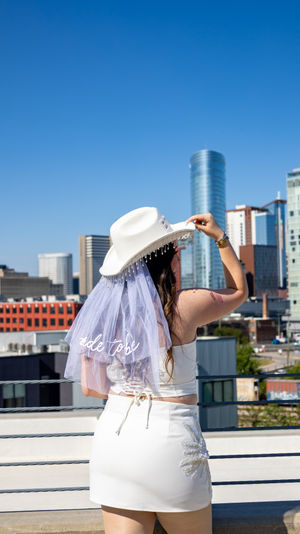 Bride-to-be in white crop top, mini skirt and beaded cowboy hat with veil reading 'bride to be,' posing on a sunny urban rooftop with modern downtown skyline and clear blue sky.