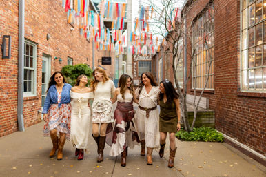 Six women laughing and arm-in-arm walking down a brick-lined downtown alley decorated with colorful hanging ribbons, wearing boho dresses and cowboy boots