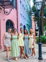 Five friends in light summer dresses smiling and posing on a sidewalk in front of pastel-colored row houses and a vintage streetlamp with palm trees in a historic tropical neighborhood