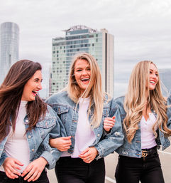 Three smiling women linked arm-in-arm on an urban rooftop, wearing denim jackets, white tops and black jeans with a downtown skyline and overcast sky in the background.