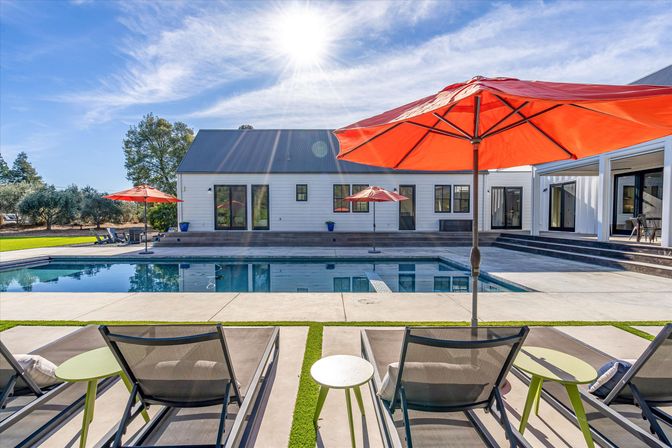 Sunny poolside scene at a modern farmhouse-style home: rectangular swimming pool, concrete patio with lounge chairs and bright orange umbrellas, landscaped lawn and mature trees under a blue sky.
