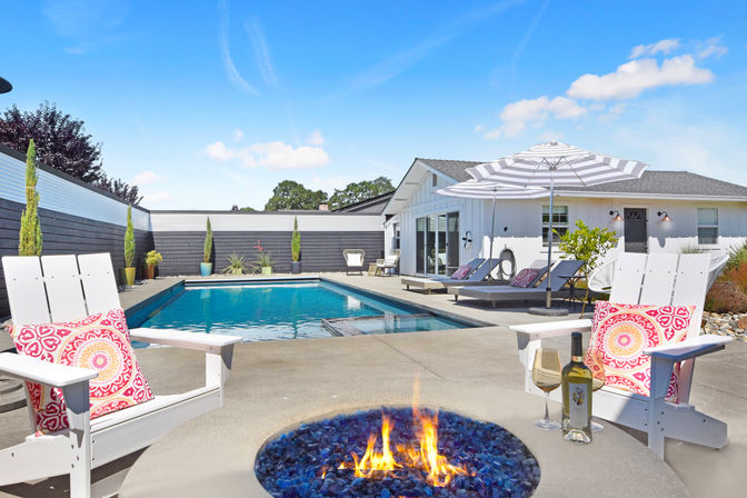 Sunny suburban backyard pool and patio — rectangular blue swimming pool with lounge chairs, striped umbrella, white Adirondack chairs with colorful pillows, glass-top fire pit and a bottle of wine for relaxed outdoor entertaining.