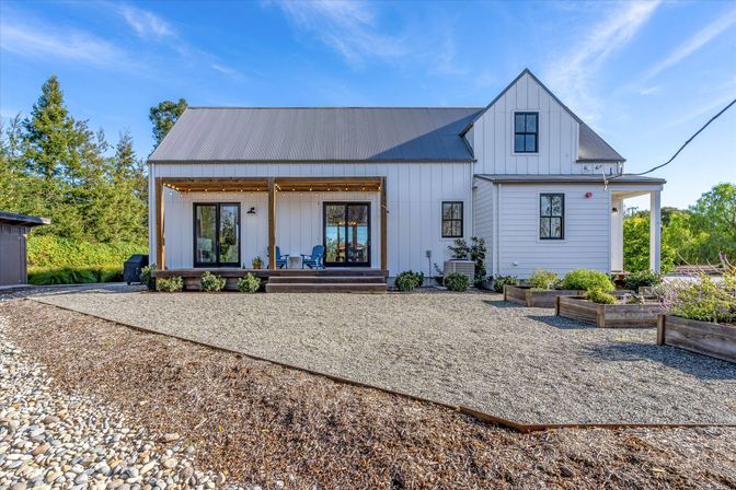 White modern farmhouse with corrugated metal roof, covered front porch with blue chairs, gravel courtyard and raised wooden planter beds under a clear blue sky