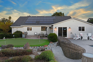 Suburban backyard of a single-story home at sunset featuring rooftop solar panels, a small putting green with flags, patio dining and grill, woven lounge sculpture, Adirondack chairs and rock-landscaped garden.