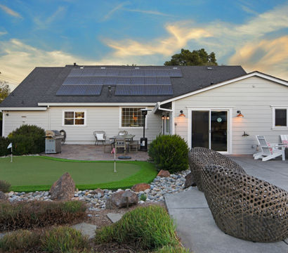 Suburban backyard of a single-story home at sunset featuring rooftop solar panels, a small putting green with flags, patio dining and grill, woven lounge sculpture, Adirondack chairs and rock-landscaped garden.