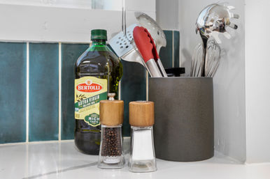 Kitchen countertop with a green bottle of extra virgin olive oil, wooden‑topped salt and pepper grinders, and a gray utensil holder with metal cooking tools against a teal tile backsplash.