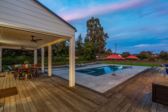 Dusk suburban backyard patio with wooden deck and ceiling fans, colorful outdoor dining, rectangular swimming pool with red umbrellas and lounge chairs