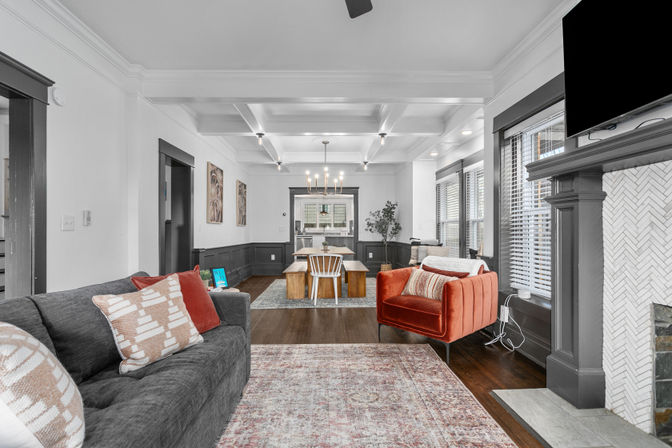 Bright open-concept living and dining room in a Craftsman-style home with coffered ceiling, dark wood trim, gray sofa, red velvet accent chair, wood floors and fireplace