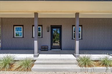 Front porch of a modern gray-shingle house with a centered black glass front door, two small windows, two square columns, concrete steps, a wall lantern and ornamental grasses flanking the walkway.