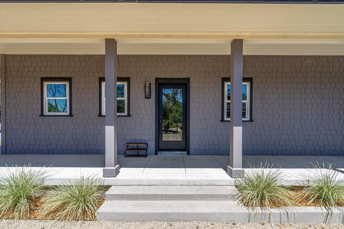 Front porch of a modern gray-shingle house with a centered black glass front door, two small windows, two square columns, concrete steps, a wall lantern and ornamental grasses flanking the walkway.