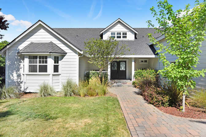 White single-story suburban house with gray shingle roof, brick paver walkway leading to black double front doors, manicured lawn, ornamental trees and shrubs under a bright blue sky
